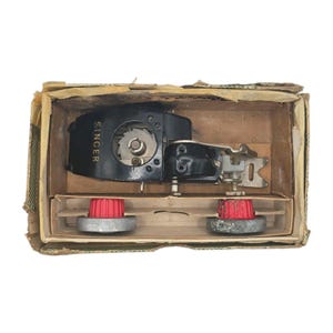 May include: A vintage black Singer sewing machine attachment with a metal gear mechanism and two red plastic adjustment knobs, housed inside a rectangular cardboard box with internal dividers and visible wear on the edges.