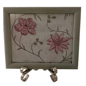 May include: A rectangular wall art piece featuring a light gray fabric background with embroidered dark pink floral patterns and thin dark green vines. The artwork is encased in a simple, smooth, light gray wooden frame and rests on a metal stand.