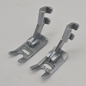 Könnte beinhalten: Zwei silberfarbene Nähmaschinen-Nähfüße aus Metall. Jedes Bauteil verfügt über einen klappbaren Schaft mit oberer Hakenbefestigung und eine flache, gegabelte Grundplatte. Die Metalloberflächen weisen ein mattes Finish auf.