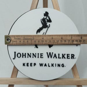 May include: A round, white sign with the Johnnie Walker logo and text. The black logo depicts a man walking with a cane. The text reads "JOHNNIE WALKER. KEEP WALKING." The sign is approximately 8 inches in diameter.