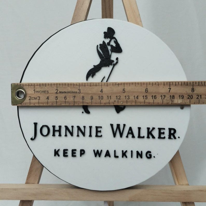 May include: A round, white sign with the Johnnie Walker logo and text. The black logo depicts a man walking with a cane. The text reads "JOHNNIE WALKER. KEEP WALKING." The sign is approximately 8 inches in diameter.