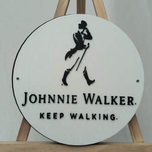 May include: A round, white sign with a black border and the Johnnie Walker logo. The logo features a walking figure with a cane. The text "JOHNNIE WALKER. KEEP WALKING." is printed in black below the logo.