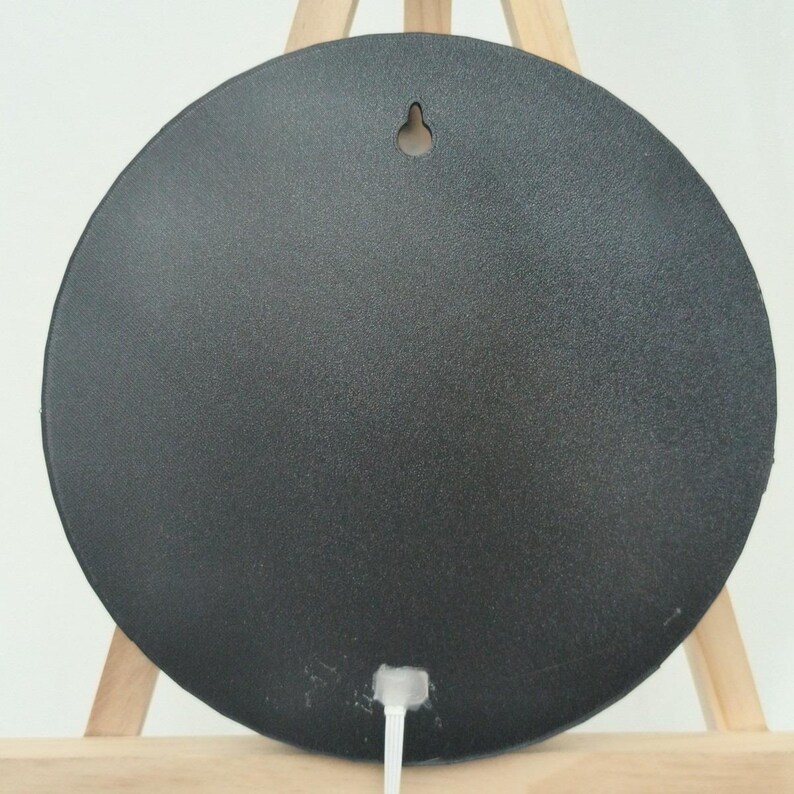 May include: A round, black object with a textured surface. It has a small hole at the top for hanging and a white cord extending from the bottom. The object is displayed on a wooden easel.
