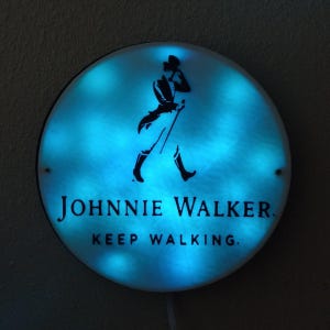 May include: A round, illuminated sign with a blue glow. The sign features the Johnnie Walker logo, a silhouette of a man walking with a cane, and the text "JOHNNIE WALKER. KEEP WALKING."
