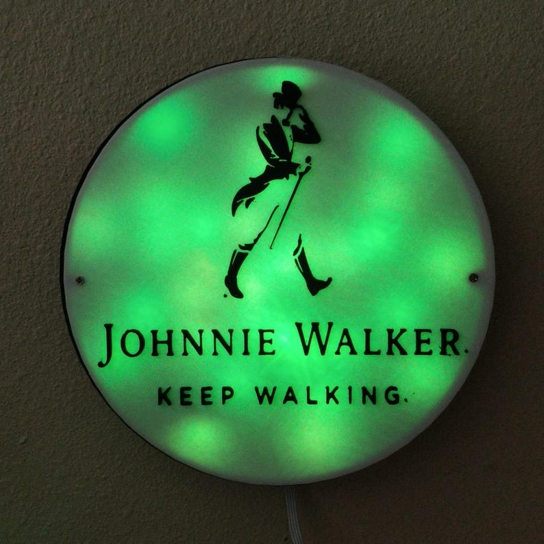 May include: Round, illuminated sign with a green glow. The sign features the Johnnie Walker logo, a silhouette of a man walking with a cane, and the text "JOHNNIE WALKER. KEEP WALKING." The sign is mounted on a neutral-colored wall.