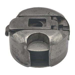 Könnte beinhalten: Eine metallene Spulenkapsel für eine Nähmaschine mit einem klappbaren Hebel an der Oberseite. An der Seite des zylindrischen Bauteils befindet sich die Prägung BOCCO 52473 U.S.A. Das Objekt hat eine silbergraue Oberfläche mit Gebrauchsspuren.