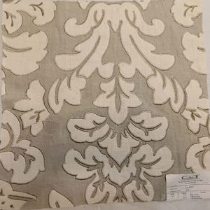 May include: A square fabric swatch featuring a large-scale, cream-colored damask pattern embroidered onto a light taupe background. The design consists of symmetrical, stylized acanthus leaves and floral motifs. A small white product label is attached.