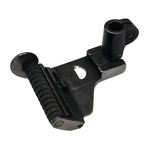 May include: A dark metallic sewing machine presser foot attachment featuring a cylindrical, textured roller wheel at the front for fabric feeding. The component includes a vertical mounting shank with a circular opening and a flat metal plate.