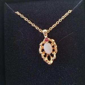 May include: A gold-colored necklace with a pendant featuring an oval, off-white stone and a small red stone. The pendant has an ornate, openwork design. The necklace is displayed in a black box.