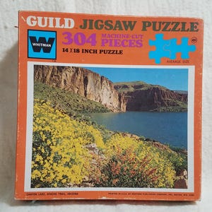 May include: Vintage Guild jigsaw puzzle box with a scenic image of Canyon Lake, Apache Trail, Arizona. The box is orange with the text "304 Pieces" and "14 x 18 inch puzzle". The puzzle features a landscape with a lake, mountains, and yellow flowers.