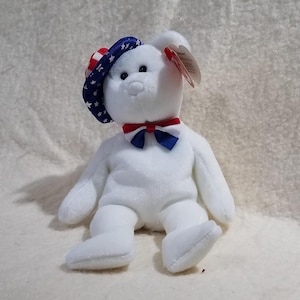 May include: A white plush bear toy featuring a blue hat with white stars and red stripes, and a red, white, and blue bowtie. The bear has black plastic eyes and a black nose, with a heart-shaped red tag attached to its ear.
