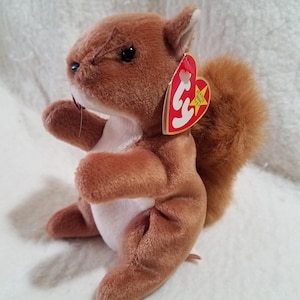 May include: A brown plush squirrel toy with a fluffy tail and a red heart-shaped tag. The squirrel is in a sitting position with its arms in front. The toy has black eyes and a white belly.
