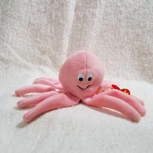 May include: A light pink plush octopus toy with a rounded head, eight soft tentacles, and two white eyes with black pupils. The toy features a small red heart-shaped tag attached to the side, displaying the word Ty in yellow lettering.