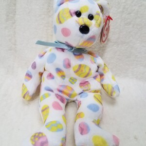 May include: A white plush bear with colorful Easter egg patterns in yellow, pink, and blue. The bear has a light blue ribbon around its neck and a red tag. A cute and cuddly Easter-themed stuffed animal.