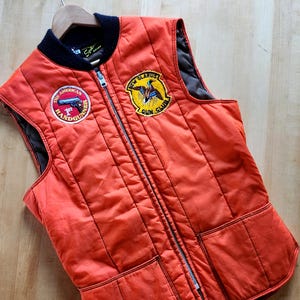 Może przedstawiać: Pomarańczowa pikowana kamizelka z czarnym kołnierzem i podszewką. Kamizelka posiada pełny zamek błyskawiczny i dwie przednie kieszenie. Naszywki to "The American Handgunner" i "New Glarus Gun Club".