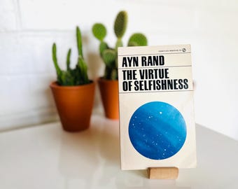 Op de afbeelding: Boek "The Virtue of Selfishness" van Ayn Rand, gepresenteerd op een houten standaard. De boekomslag heeft een blauw cirkelontwerp. Twee cactussen in potten staan op de achtergrond. Het boek staat op een wit oppervlak.