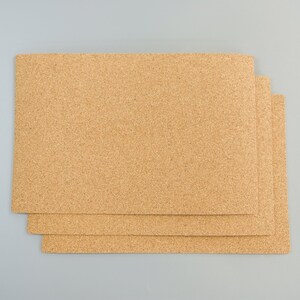 May include: Three rectangular cork placemats with a natural, light brown color. The placemats are stacked on top of each other, with the top one slightly offset to reveal the others. The cork material has a textured surface.