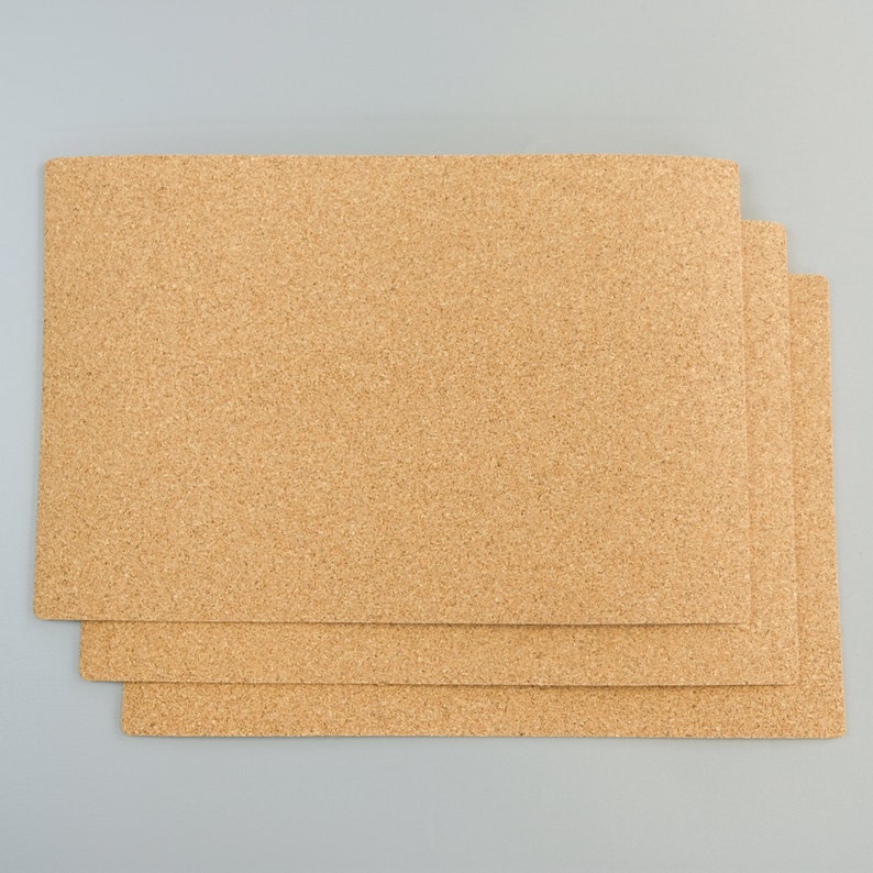 May include: Three rectangular cork placemats with a natural, light brown color. The placemats are stacked on top of each other, with the top one slightly offset to reveal the others. The cork material has a textured surface.
