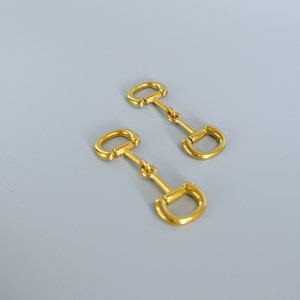 Può includere: Due orecchini in metallo color oro con design a morso di cavallo. Ogni orecchino presenta un piccolo anello nella parte superiore, collegato a una barra centrale e a un anello più grande a forma di D nella parte inferiore. Gli orecchini sono esposti su uno sfondo grigio chiaro.