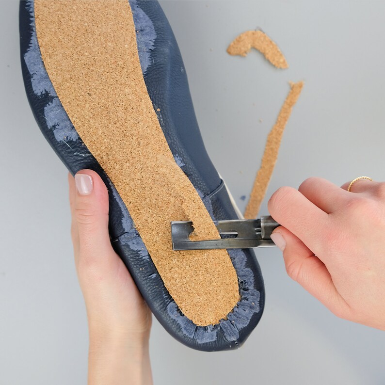 May include: A close-up of a shoe sole being trimmed with a blade. The shoe is navy blue leather with a cork insole. The blade is cutting away excess cork material. The background is a light gray surface.