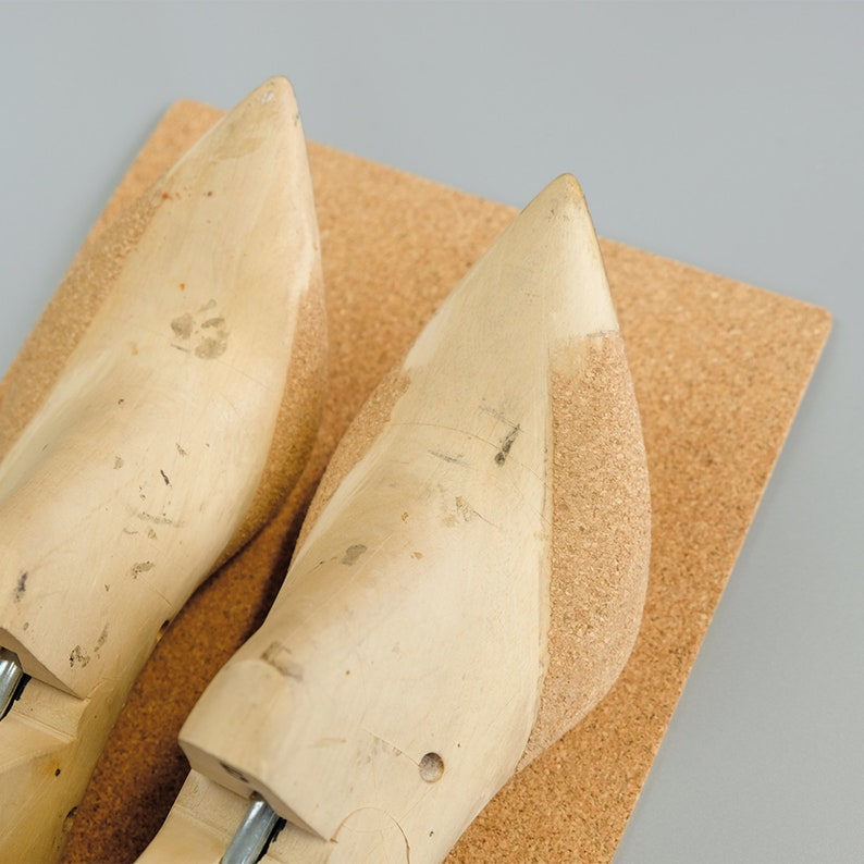 May include: Two wooden shoe forms with a cork base. The forms are light-colored with a pointed toe and show signs of wear. The cork base is rectangular and light brown. The background is a solid gray.