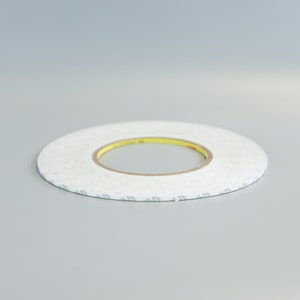Puede incluir: Un rollo de cinta adhesiva de doble cara blanca con un núcleo amarillo. La cinta tiene un diseño estampado en un lado.