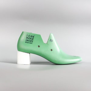 May include: A green plastic shoe last with a white heel. The last has the text "I CAN MAKE SHOES RECYCLED PLASTIC" printed on the side. The size 39 is printed on the top of the shoe.