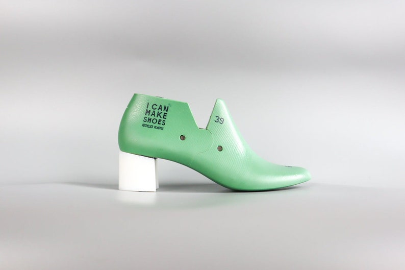 May include: A green plastic shoe last with a white heel. The last has the text "I CAN MAKE SHOES RECYCLED PLASTIC" printed on the side. The size 39 is printed on the top of the shoe.