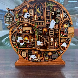 May include: A wooden, multi-layered, illuminated diorama shaped like a library. It features white ghosts, a black cat, pumpkins, and bookshelves. Text includes 'The Ghostly Library', 'Read More Books', 'Quiet Please', 'Stories Live Here', and 'Cozy Readers Club'.