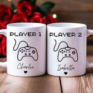 May include: Two white ceramic mugs with black text and graphics. One mug says "PLAYER 1" above a controller graphic and the name "Charlie" below. The other mug says "PLAYER 2" above a controller graphic and the name "Isabella" below.