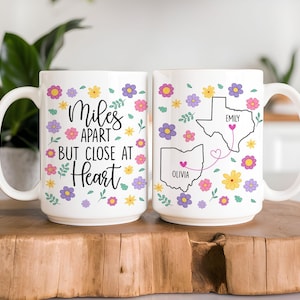 Könnte beinhalten: Weiße Keramiktasse mit dem Text "Miles Apart But Close at Heart", umgeben von bunten Blumen. Die zweite Tasse zeigt Umrisse von Bundesstaaten mit Namen und Herzen, die eine Fernbeziehung symbolisieren.
