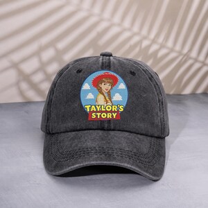 May include: A charcoal gray baseball cap featuring a circular patch on the front. The patch displays an illustration of a person wearing a red cowboy hat against a blue sky with clouds, with the yellow and red text "TAYLOR'S STORY" below.