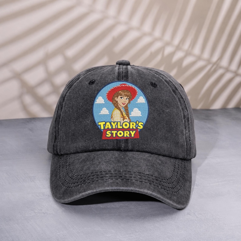 May include: A charcoal gray baseball cap featuring a circular patch on the front. The patch displays an illustration of a person wearing a red cowboy hat against a blue sky with clouds, with the yellow and red text "TAYLOR'S STORY" below.