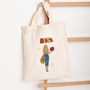 May include: A natural-coloured canvas tote bag featuring a graphic print of a person with long blonde hair, wearing blue jeans and cowboy boots, holding a guitar and a red hat. Above the figure, the letters TS are printed in yellow on a red shape.