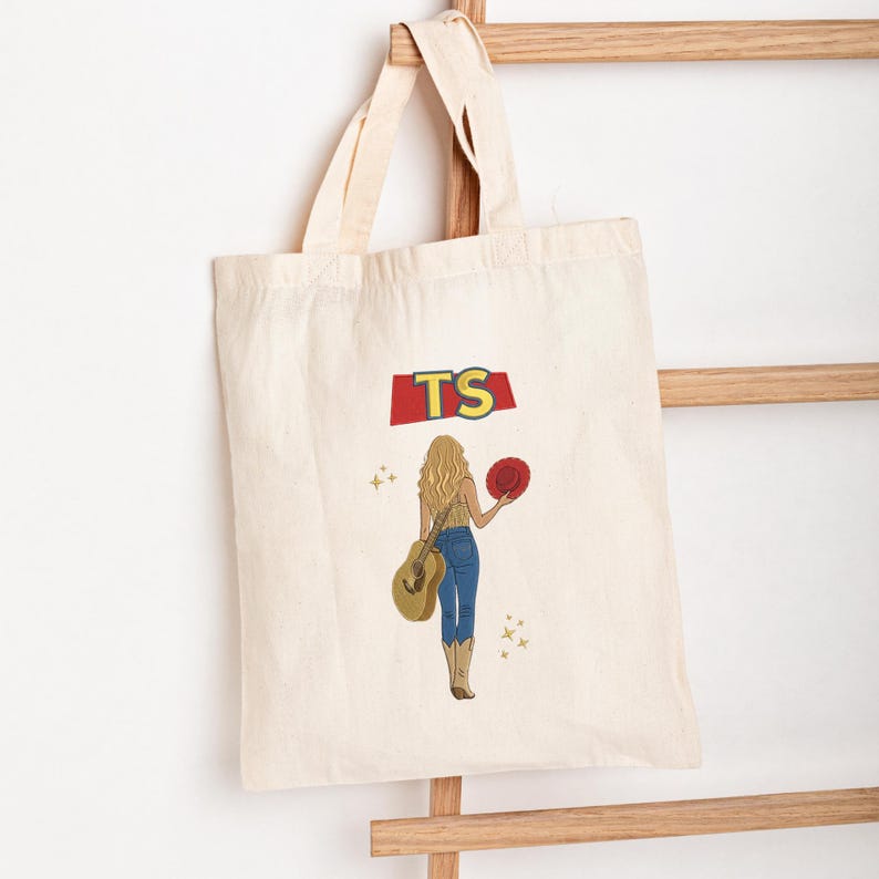 May include: A natural-coloured canvas tote bag featuring a graphic print of a person with long blonde hair, wearing blue jeans and cowboy boots, holding a guitar and a red hat. Above the figure, the letters TS are printed in yellow on a red shape.