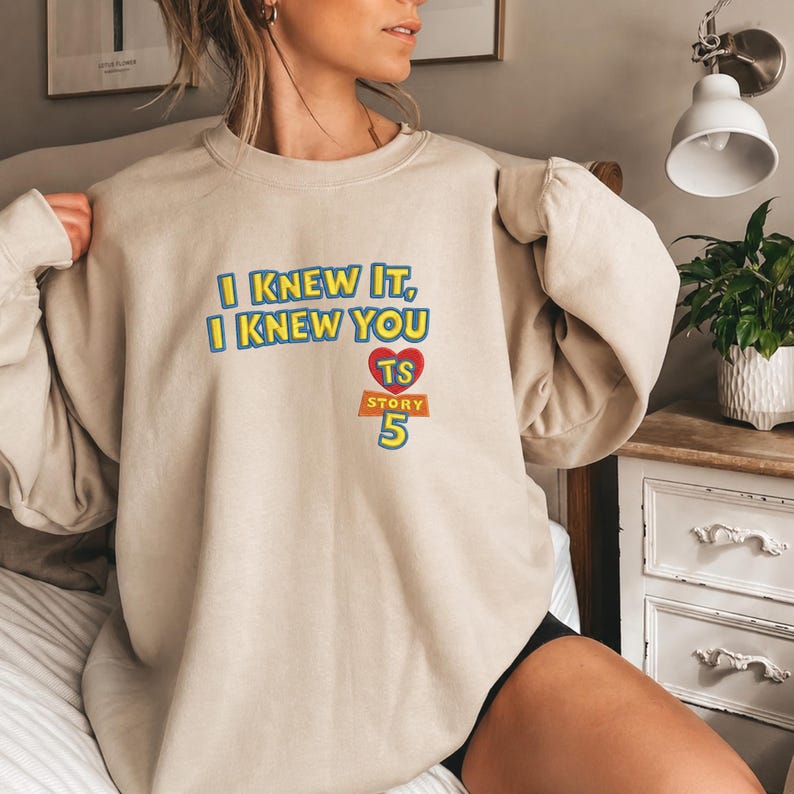 May include: A beige crewneck sweatshirt featuring the blue text I KNEW IT, I KNEW YOU, a red heart with the letters TS, and the text STORY 5 in yellow with a blue outline. The design is centered on the chest of the long-sleeved garment.