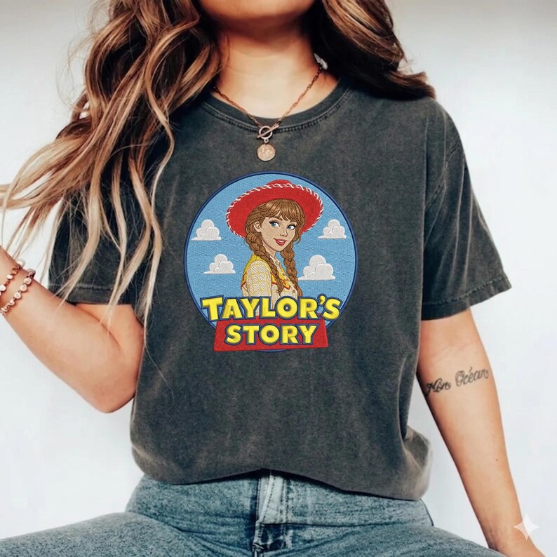 May include: A charcoal gray short-sleeve t-shirt featuring a circular graphic of a woman with braided hair wearing a red cowboy hat. The design includes a blue sky background with white clouds and the yellow and red text Taylor's Story.
