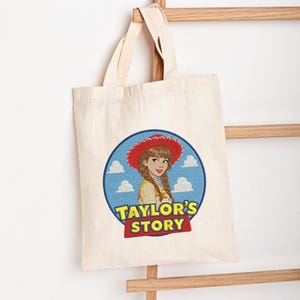 May include: A natural-colored canvas tote bag featuring a circular graphic of a person with braided hair wearing a red cowboy hat. The design includes a blue sky with white clouds and the yellow and red text "TAYLOR'S STORY" at the bottom.