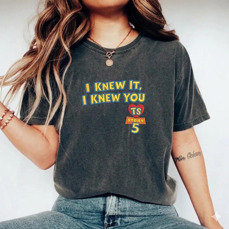 May include: A charcoal gray short-sleeve t-shirt featuring the yellow text I KNEW IT, I KNEW YOU, a red heart with the letters TS, and the orange text STORY 5. The shirt has a crew neckline and a relaxed fit, made of a soft, washed-out cotton fabric.