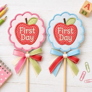 May include: Two round scalloped fabric signs on wooden sticks, one with pink gingham and one with blue gingham. Each features a red apple appliqué with the text First Day in white embroidery, accented by a green leaf and multi-colored ribbons.