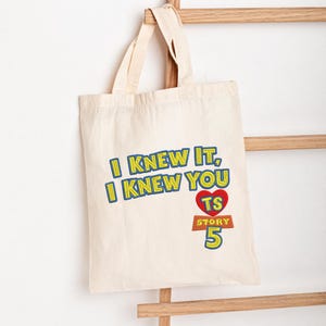 May include: A natural-colored canvas tote bag featuring embroidered text that reads I KNEW IT, I KNEW YOU, with a red heart containing the letters TS above the word STORY and the number 5 in blue, yellow, and red thread on the front.