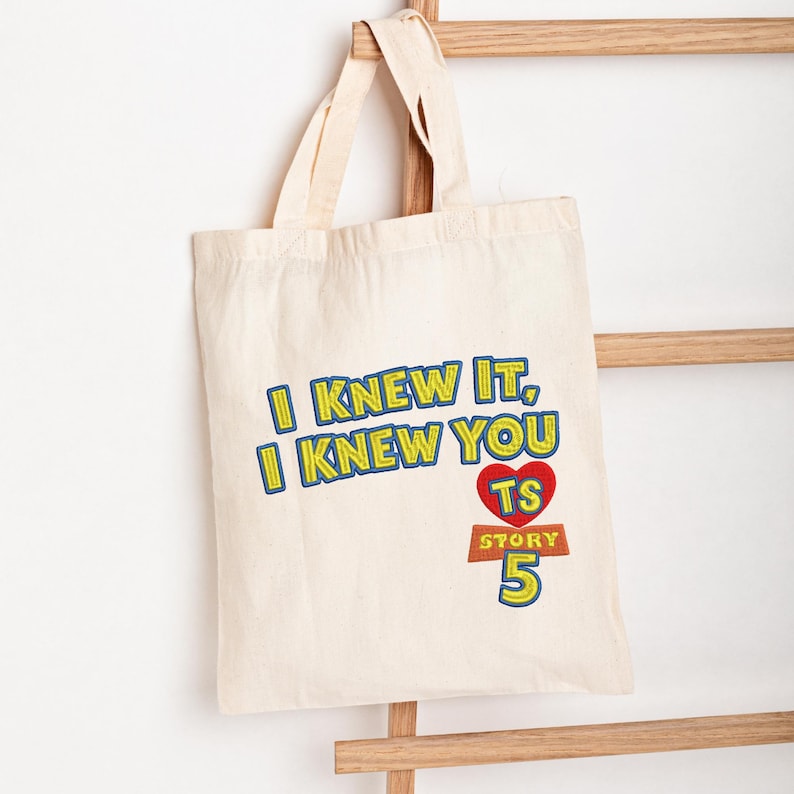 May include: A natural-colored canvas tote bag featuring embroidered text that reads I KNEW IT, I KNEW YOU, with a red heart containing the letters TS above the word STORY and the number 5 in blue, yellow, and red thread on the front.