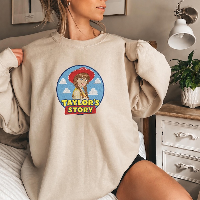 May include: A beige crewneck sweatshirt featuring a circular graphic on the chest. The graphic depicts a cartoon character with braided hair wearing a red cowboy hat against a blue sky with white clouds. The text "TAYLOR'S STORY" is printed below.