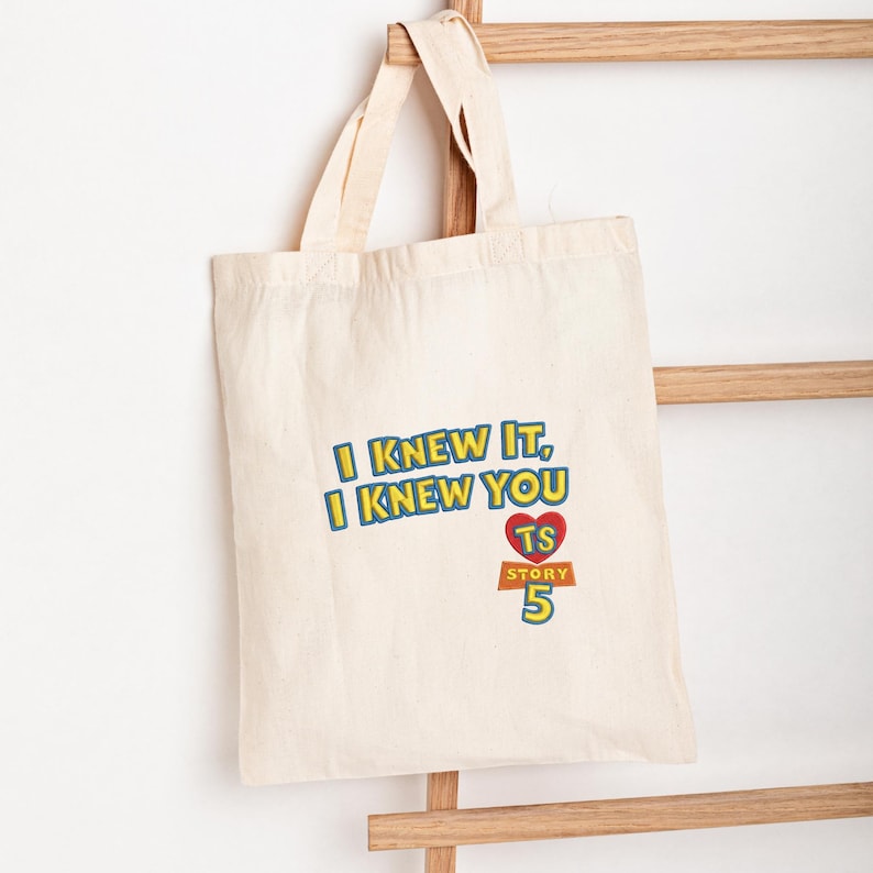 May include: A natural-colored canvas tote bag with two handles. The front features the blue and yellow text "I KNEW IT, I KNEW YOU" above a red heart containing the letters "TS" and the text "STORY 5" in yellow and orange lettering.