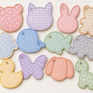 Puede incluir: Una colección de doce parches de tela con diversas formas de animales e insectos, incluyendo oso, gato, conejo, ballena, pez, elefante, pato, mariposa y dinosaurio. Cada parche presenta un patrón pastel único y un pequeño orificio circular.