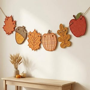 May include: A decorative garland featuring six hanging ornaments shaped like autumn leaves, an acorn, a pumpkin, and an apple. Each piece is crafted with orange, yellow, and red fabric patterns and is strung together on a thin, light-colored twine rope.