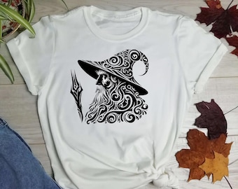 May include: White t-shirt featuring a black graphic of a wizard with a long beard, hat, and staff. The wizard design is detailed with swirling patterns. The shirt is made of a soft material and has short sleeves.