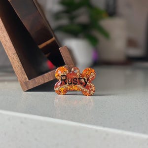 May include: A bone-shaped pet tag with the name "Rusty" in black lettering. The tag features a mix of orange, gold, and brown glitter, creating a sparkling effect. The tag is on a light-colored surface.