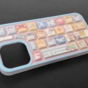 May include: A light blue phone case with a keyboard design. The case features a series of colorful, square keys with cartoon character illustrations. The keys are in shades of yellow, pink, blue, and white. The case has a rounded edge and a cutout for the phone's camera.