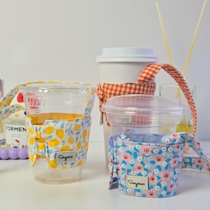 May include: Three fabric cup sleeves with handles, each featuring a small rectangular tag labeled Soyna. One sleeve has a yellow lemon print, one has an orange and white gingham pattern, and one has a light blue floral print with pink flowers.